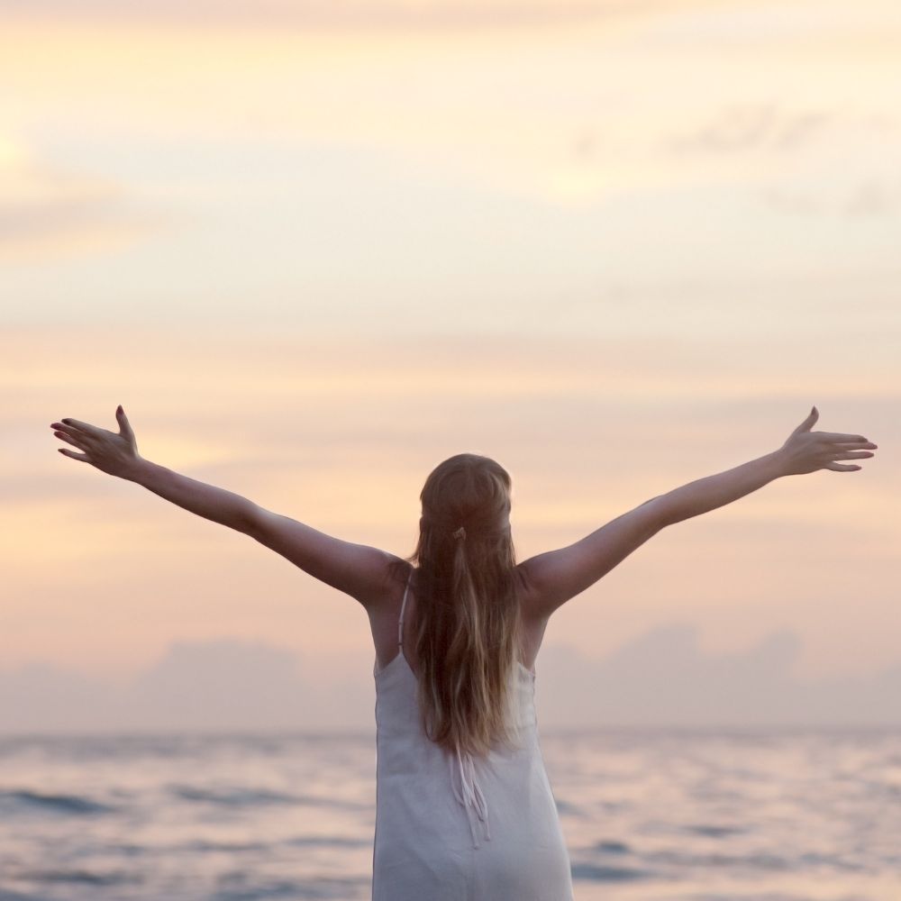 a woman wit arms stretched upwards looking out to sea